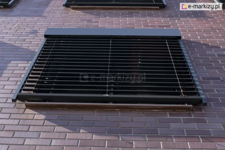 Zewnętrzna żaluzja fasadowa Z90 Cube w kolorze ciemnego antracytu zamontowana na elewacji wykonanej z klinkierowych cegieł w odcieniach brązu i fioletu. Żaluzja jest opuszczona i zasłania okno, a jej lamele są ustawione poziomo, pozwalając na częściowy dopływ światła do wnętrza. Konstrukcja zawiera prowadnice boczne oraz górną kasetę osłonową dopasowaną kolorystycznie do lameli. Poniżej dolnej krawędzi żaluzji widoczny jest zewnętrzny parapet. Zdjęcie wykonane pod kątem z dołu, ukazujące detal montażu systemu osłonowego na ceglanej ścianie budynku.