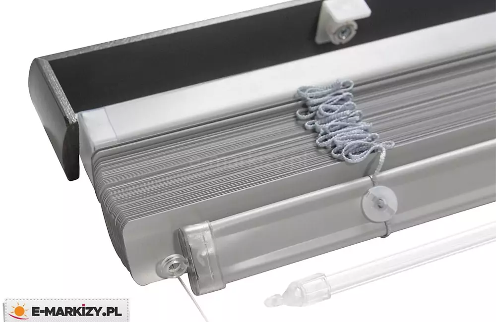 Zdjęcie prezentuje techniczne detale konstrukcyjne, jakimi wyróżniają się żaluzje poziome aluminiowe w kolorze srebrnym lub jasnoszarym. Widok z bliska ukazuje górną rynnę montażową oraz pakiet złożonych, ściśle przylegających do siebie lameli. Górna szyna wyposażona została w dodatkową maskownicę drewnianą o kanciasstym kształcie, wykończoną w kolorze ciemnego antracytu lub czerni, co stanowi kontrast dla jasnych listewek. Wewnątrz rynny górnej widoczny jest biały element mechanizmu mocującego. Z pakietu zebranych lameli wyłania się szara, zapleciona drabinka sznurkowa, która odpowiada za utrzymanie poziomego układu żaluzji po ich rozłożeniu. Na spodzie widoczna jest dolna belka obciążająca, zakończona przezroczystą, plastikową zaślepką, przez którą przechodzi cienka żyłka prowadzenia bocznego. W dolnej części kadru znajduje się również fragment przezroczystego, akrylowego pokrętła (patyka), służącego do regulacji kąta nachylenia lameli. Całość mechanizmu jest przedstawiona na czystym, białym tle, co pozwala na dokładne przyjrzenie się komponentom takim jak otwory w lamelach, przez które przechodzą sznurki, oraz precyzja spasowania poszczególnych elementów osłony.