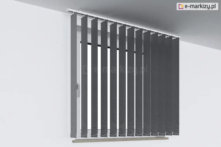 Zdjęcie przedstawia nowoczesne żaluzje pionowe vertikal w kolorze ciemnoszarym lub antracytowym, zamontowane na białej ścianie nad oknem. System składa się z wielu wąskich, pionowych pasów tkaniny, które są podwieszone do białej szyny montażowej przymocowanej bezpośrednio pod sufitem. Lamele są ustawione prostopadle do płaszczyzny okna w pozycji otwartej, co pozwala na wgląd do wnętrza wnęki okiennej, gdzie widoczna jest biała rama okna z klamką. Żaluzje pionowe vertikal osłaniają całą szerokość otworu okiennego, sięgając nieco poniżej linii wąskiego, drewnianego parapetu o jasnym odcieniu. Na dole każdego pasa materiału znajduje się biały mechanizm obciążający, a wszystkie lamele są połączone ze sobą subtelnym, białym łańcuszkiem stabilizującym, który układa się w równe pętle. Minimalistyczna aranżacja na tle gładkich, jasnych ścian podkreśla kontrast między ciemnym materiałem osłony a jasnym otoczeniem, prezentując produkt w sposób techniczny i przejrzysty.