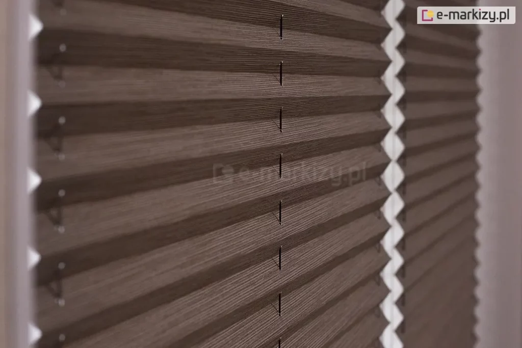 Zbliżenie na materiałową żaluzję plisowaną w ciemnobrązowym, stonowanym odcieniu o wyraźnej, tekstylnej strukturze przypominającej splot tkaniny. Zdjęcie prezentuje detal harmonijkowej konstrukcji osłony okiennej, ukazując regularne, ostre zagięcia materiału tworzące powtarzalny wzór poziomych linii i cieni. Przez środek każdej plisy przechodzą pionowo poprowadzone, cienkie sznurki prowadzące, które stabilizują konstrukcję i umożliwiają jej precyzyjne składanie oraz rozkładanie. Żaluzja plisowana jest zamontowana w białej ramie okiennej, która jest widoczna po lewej stronie kadru oraz w głębi, gdzie białe elementy montażowe oddzielają od siebie dwa panele materiału. Światło padające na żaluzję podkreśla trójwymiarowość fałd oraz matowe wykończenie tkaniny, tworząc delikatne przejścia tonalne między oświetlonymi krawędziami a zagłębieniami plis. Tło po prawej stronie jest lekko rozmyte, co skupia uwagę na precyzji wykonania i fakturze produktu w pierwszym planie.