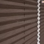 Zbliżenie na materiałową żaluzję plisowaną w ciemnobrązowym, stonowanym odcieniu o wyraźnej, tekstylnej strukturze przypominającej splot tkaniny. Zdjęcie prezentuje detal harmonijkowej konstrukcji osłony okiennej, ukazując regularne, ostre zagięcia materiału tworzące powtarzalny wzór poziomych linii i cieni. Przez środek każdej plisy przechodzą pionowo poprowadzone, cienkie sznurki prowadzące, które stabilizują konstrukcję i umożliwiają jej precyzyjne składanie oraz rozkładanie. Żaluzja plisowana jest zamontowana w białej ramie okiennej, która jest widoczna po lewej stronie kadru oraz w głębi, gdzie białe elementy montażowe oddzielają od siebie dwa panele materiału. Światło padające na żaluzję podkreśla trójwymiarowość fałd oraz matowe wykończenie tkaniny, tworząc delikatne przejścia tonalne między oświetlonymi krawędziami a zagłębieniami plis. Tło po prawej stronie jest lekko rozmyte, co skupia uwagę na precyzji wykonania i fakturze produktu w pierwszym planie.