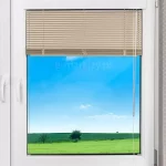 Zdjęcie prezentuje białe okno z tworzywa sztucznego z zamontowaną osłoną okienną, którą są żaluzje poziome aluminiowe w kolorze beżowym lub piaskowym. Żaluzje są częściowo podciągnięte do góry, zasłaniając górną jedną trzecią powierzchni szyby, podczas gdy dolna część okna pozostaje odsłonięta, ukazując widok na zielony krajobraz z polami i błękitnym niebem. Lamele żaluzji są ułożone równolegle i ściśle przylegają do siebie w górnej części, tworząc zwartą strukturę. Przez środek oraz po bokach żaluzji przechodzą cienkie, pionowe sznurki drabinki, które stabilizują konstrukcję i umożliwiają sterowanie mechanizmem. Po prawej stronie zwisają dwa przezroczyste sznurki służące do podnoszenia i opuszczania osłony, zakończone niewielkim elementem dociążającym. Żaluzje są zamontowane bezpośrednio na ramie skrzydła okiennego w świetle szyby. Górna rynna żaluzji jest koloru białego, co spójnie komponuje się z ramą okna. Rama okienna posiada widoczne zawiasy po prawej stronie oraz białą klamkę po lewej stronie skrzydła. Ściana wokół okna wykończona jest tapetą w delikatny, geometryczny wzór składający się z małych jasnych kółek na szarym tle. Światło padające na żaluzje podkreśla ich metaliczną, gładką fakturę oraz precyzyjne wykonanie poziomych listewek.