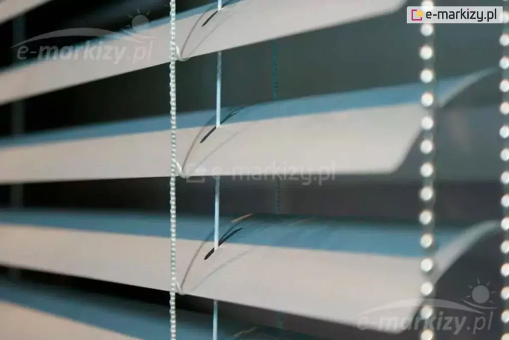 Zbliżenie na poziome żaluzje aluminiowe 25 mm w kolorze jasnoszarym lub srebrnym, zamontowane we wnętrzu pomieszczenia. Konstrukcja składa się z równolegle ułożonych, lekko wygiętych lameli o szerokości dwudziestu pięciu milimetrów każda, które są ustawione pod kątem, tworząc efekt światłocienia na ich powierzchni. Przez środek lameli przechodzą pionowe, białe sznurki drabinki oraz cieńsza żyłka prowadzenia bocznego, która stabilizuje osłonę okienną. Widoczne są precyzyjnie wycięte otwory technologiczne w kształcie wydłużonych elips, przez które przewleczone są elementy sterujące konstrukcją. Po prawej stronie kadru znajduje się pionowy łańcuszek operacyjny wykonany z drobnych, gęsto rozmieszczonych koralików z tworzywa sztucznego, służący do regulacji kąta nachylenia listew. Tło za żaluzjami pozostaje nieostre i ciemne, co podkreśla detale techniczne osłony, takie jak faktura aluminium, splot sznurków oraz mechanizm podtrzymujący lamele w równych odstępach.