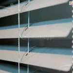 Zbliżenie na poziome żaluzje aluminiowe 25 mm w kolorze jasnoszarym lub srebrnym, zamontowane we wnętrzu pomieszczenia. Konstrukcja składa się z równolegle ułożonych, lekko wygiętych lameli o szerokości dwudziestu pięciu milimetrów każda, które są ustawione pod kątem, tworząc efekt światłocienia na ich powierzchni. Przez środek lameli przechodzą pionowe, białe sznurki drabinki oraz cieńsza żyłka prowadzenia bocznego, która stabilizuje osłonę okienną. Widoczne są precyzyjnie wycięte otwory technologiczne w kształcie wydłużonych elips, przez które przewleczone są elementy sterujące konstrukcją. Po prawej stronie kadru znajduje się pionowy łańcuszek operacyjny wykonany z drobnych, gęsto rozmieszczonych koralików z tworzywa sztucznego, służący do regulacji kąta nachylenia listew. Tło za żaluzjami pozostaje nieostre i ciemne, co podkreśla detale techniczne osłony, takie jak faktura aluminium, splot sznurków oraz mechanizm podtrzymujący lamele w równych odstępach.