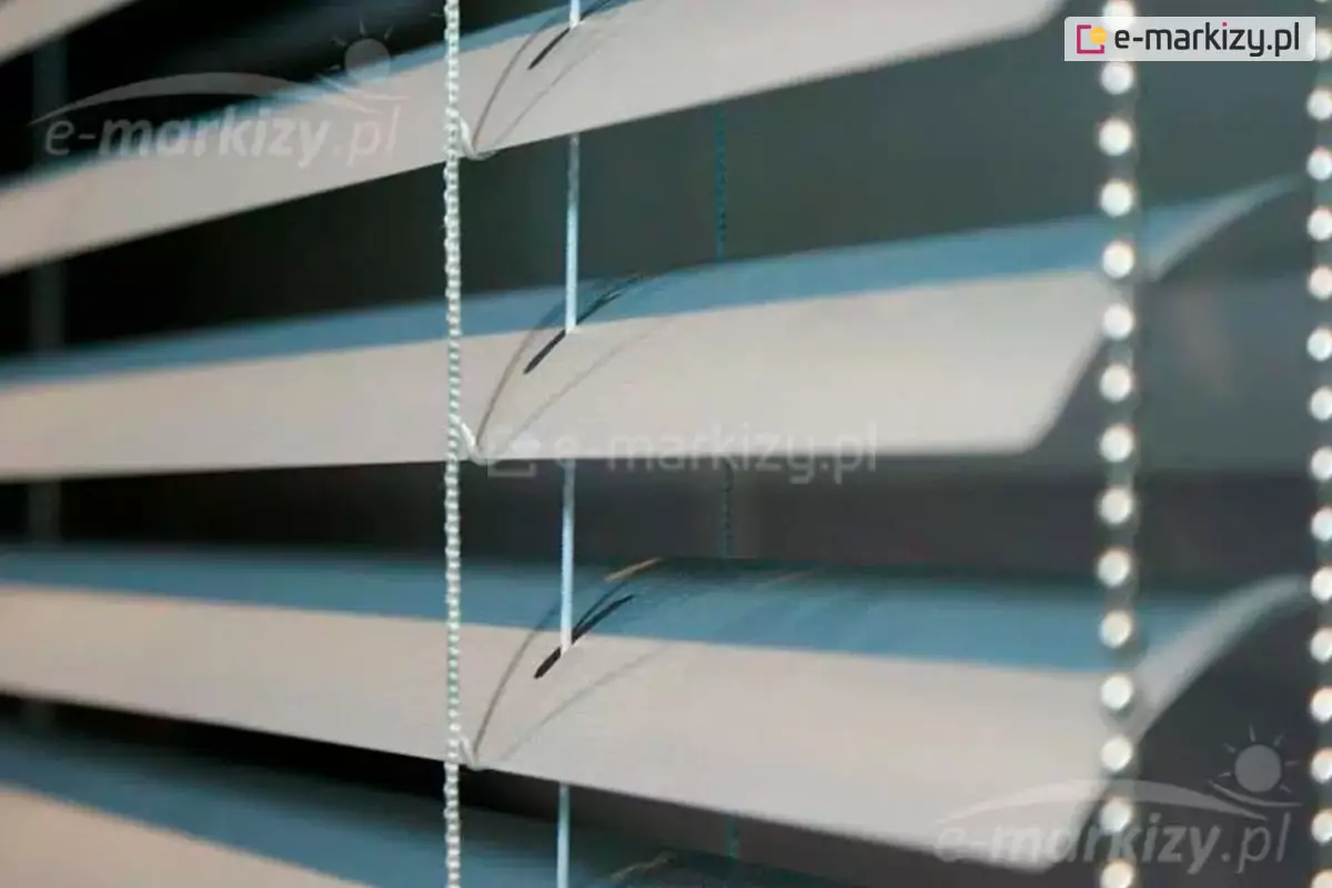 Zbliżenie na poziome żaluzje aluminiowe 25 mm w kolorze jasnoszarym lub srebrnym, zamontowane we wnętrzu pomieszczenia. Konstrukcja składa się z równolegle ułożonych, lekko wygiętych lameli o szerokości dwudziestu pięciu milimetrów każda, które są ustawione pod kątem, tworząc efekt światłocienia na ich powierzchni. Przez środek lameli przechodzą pionowe, białe sznurki drabinki oraz cieńsza żyłka prowadzenia bocznego, która stabilizuje osłonę okienną. Widoczne są precyzyjnie wycięte otwory technologiczne w kształcie wydłużonych elips, przez które przewleczone są elementy sterujące konstrukcją. Po prawej stronie kadru znajduje się pionowy łańcuszek operacyjny wykonany z drobnych, gęsto rozmieszczonych koralików z tworzywa sztucznego, służący do regulacji kąta nachylenia listew. Tło za żaluzjami pozostaje nieostre i ciemne, co podkreśla detale techniczne osłony, takie jak faktura aluminium, splot sznurków oraz mechanizm podtrzymujący lamele w równych odstępach.