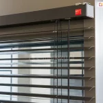 Górna część żaluzji fasadowej C50 Slim Selt z widoczną kwadratową kasetą oraz wyprowadzonym z niej systemem sterowania i drabinką sznurkową pomiędzy lamelami w odcieniu grafitu. Kaseta zawieszona na ścianie za pomocą specjalnych uchwytów, które pozostają prawie niewidoczne.