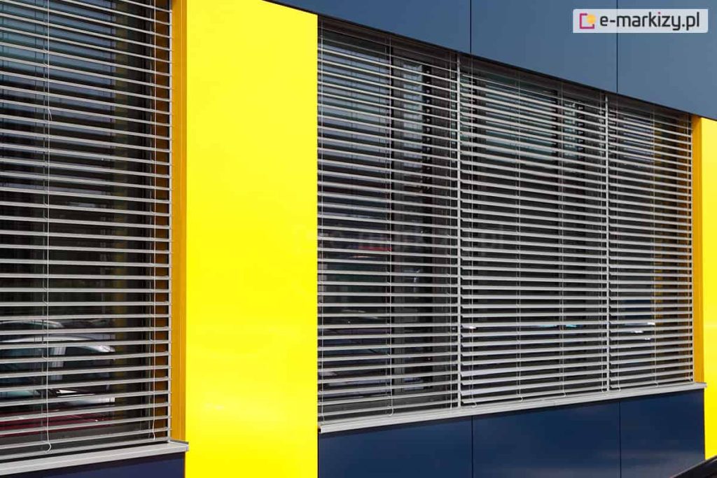 Fragment nowoczesnej fasady budynku z dużymi przeszkleniami, zasłoniętymi zewnętrznymi żaluzjami fasadowymi. Żaluzje mają poziome aluminiowe lamele w kolorze srebrnym, ustawione równolegle i prowadzone za pomocą pionowych linek. Lamele są uchylone, co umożliwia częściowe przenikanie światła. Elewacja budynku wykończona jest kolorowymi panelami elewacyjnymi – intensywnie żółtymi po bokach i ciemnogranatowymi (prawie czarnymi) powyżej i poniżej okien. Szyby są lekko refleksyjne, odbijają otoczenie i światło.