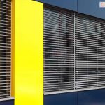 Fragment nowoczesnej fasady budynku z dużymi przeszkleniami, zasłoniętymi zewnętrznymi żaluzjami fasadowymi. Żaluzje mają poziome aluminiowe lamele w kolorze srebrnym, ustawione równolegle i prowadzone za pomocą pionowych linek. Lamele są uchylone, co umożliwia częściowe przenikanie światła. Elewacja budynku wykończona jest kolorowymi panelami elewacyjnymi – intensywnie żółtymi po bokach i ciemnogranatowymi (prawie czarnymi) powyżej i poniżej okien. Szyby są lekko refleksyjne, odbijają otoczenie i światło.