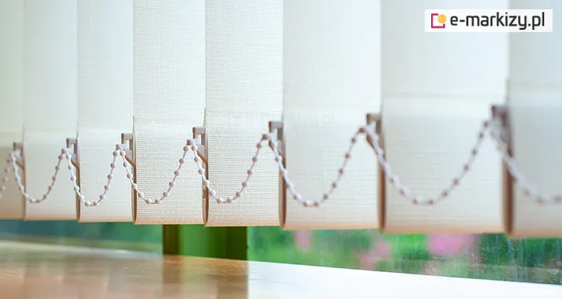 Zdjęcie prezentuje zbliżenie na dolną krawędź żaluzji pionowych w kolorze kremowym lub bardzo jasnym beżu, które znajdują się tuż nad gładkim, drewnianym blatem lub parapetem. Poszczególne pasy materiału mają wyraźną, tekstylną strukturę o drobnym splocie, a ich dolne części są wywinięte, tworząc kieszenie na obciążniki stabilizujące. Po bokach każdej lameli znajdują się plastikowe uchwyty, przez które poprowadzono biały łańcuszek kulkowy, łączący wszystkie elementy w jeden system. Łańcuszek układa się w powtarzalne, miękkie łuki, co jest charakterystyczne dla mechanizmu żaluzji pionowych. W tle, za przesłoną, widoczne jest okno, przez które prześwituje zieleń ogrodu z różowymi akcentami kwiatów, jednak obraz ten jest mocno rozmyty, co skupia uwagę na detalach technicznych osłony. Światło dzienne wpadające z zewnątrz delikatnie rozświetla krawędzie lameli, podkreślając ich pionowy układ.