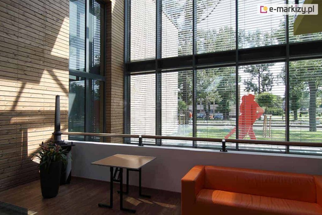 Żaluzje fasadowe zamocowane na dużej szklanej ścianie - łączone modułowo na szerokość oraz ustawione jedne nad drugimi. Za oknem widoczna tańcząca para oraz parki z drzewami i trawnikiem. Przed oknem wewnątrz budynku stolik i kanapa w kolorze pomarańczowym oraz roślina w dużej ciemnej donicy.