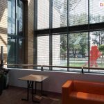 Żaluzje fasadowe zamocowane na dużej szklanej ścianie - łączone modułowo na szerokość oraz ustawione jedne nad drugimi. Za oknem widoczna tańcząca para oraz parki z drzewami i trawnikiem. Przed oknem wewnątrz budynku stolik i kanapa w kolorze pomarańczowym oraz roślina w dużej ciemnej donicy.