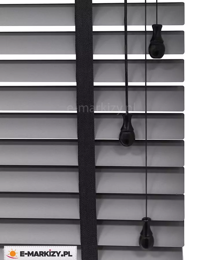 Zdjęcie prezentuje szczegółowe zbliżenie na żaluzje aluminiowe 50 mm w eleganckim, matowym odcieniu szarości lub antracytu. Szerokie, poziome lamele o szerokości pięćdziesięciu milimetrów są ułożone równolegle, tworząc rytmiczną strukturę z wyraźnymi odstępami, przez które prześwituje białe tło. Kluczowym elementem dekoracyjnym i technicznym jest pionowa, szeroka drabinka taśmowa w kolorze głębokiej czerni, wykonana z gęsto tkanej tkaniny o widocznym splocie, która podtrzymuje lamele. Po prawej stronie kadru znajdują się trzy czarne sznurki operacyjne o różnej długości, z których każdy zakończony jest ergonomicznym, dekoracyjnym obciążnikiem w kształcie łezki wykonanym z czarnego tworzywa. Sznurki te służą do sterowania mechanizmem podnoszenia oraz kątem nachylenia listew. Kompozycja podkreśla nowoczesny design oraz kontrastowe połączenie kolorystyczne między szarymi lamelami a czarnym osprzętem, co jest charakterystyczne dla systemów, w których wykorzystywana jest żaluzja aluminiowa 50 mm.
