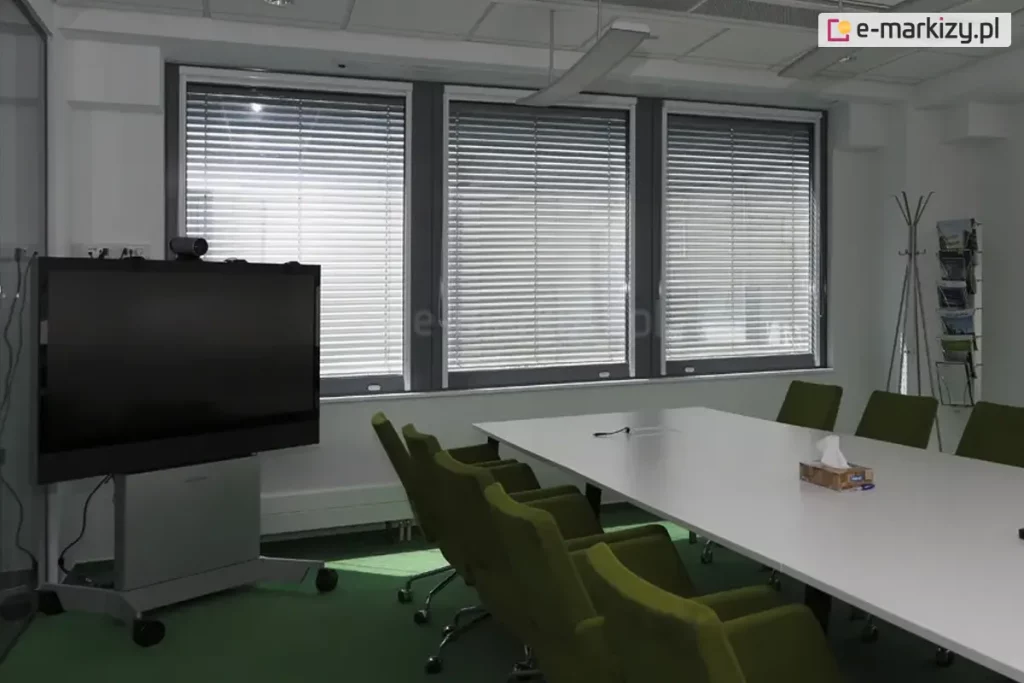 Widok na nowoczesną salę konferencyjną wyposażoną w system żaluzji fasadowych C50 Slim. Trzy duże okna o ciemnych profilach zostały osłonięte srebrnymi lamelami aluminiowymi, które ustawiono w pozycji zamkniętej, co pozwala na skuteczne zaciemnienie pomieszczenia i redukcję odblasków podczas prezentacji multimedialnych. Wnętrze charakteryzuje się profesjonalnym wyposażeniem: centralnie umieszczono długi, biały stół konferencyjny z ergonomicznymi fotelami w kolorze oliwkowej zieleni. Po lewej stronie widoczny jest wielkoformatowy monitor interaktywny na mobilnej podstawie, a po prawej minimalistyczny stojak na broszury. Jasne ściany i zielona wykładzina podłogowa w połączeniu z precyzyjnie dopasowanymi żaluzjami biurowymi tworzą funkcjonalną i estetyczną przestrzeń do pracy grupowej, zapewniając pełną kontrolę nad natężeniem światła wpadającego z zewnątrz.