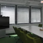 Widok na nowoczesną salę konferencyjną wyposażoną w system żaluzji fasadowych C50 Slim. Trzy duże okna o ciemnych profilach zostały osłonięte srebrnymi lamelami aluminiowymi, które ustawiono w pozycji zamkniętej, co pozwala na skuteczne zaciemnienie pomieszczenia i redukcję odblasków podczas prezentacji multimedialnych. Wnętrze charakteryzuje się profesjonalnym wyposażeniem: centralnie umieszczono długi, biały stół konferencyjny z ergonomicznymi fotelami w kolorze oliwkowej zieleni. Po lewej stronie widoczny jest wielkoformatowy monitor interaktywny na mobilnej podstawie, a po prawej minimalistyczny stojak na broszury. Jasne ściany i zielona wykładzina podłogowa w połączeniu z precyzyjnie dopasowanymi żaluzjami biurowymi tworzą funkcjonalną i estetyczną przestrzeń do pracy grupowej, zapewniając pełną kontrolę nad natężeniem światła wpadającego z zewnątrz.