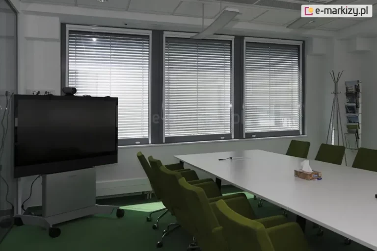 Widok na nowoczesną salę konferencyjną wyposażoną w system żaluzji fasadowych C50 Slim. Trzy duże okna o ciemnych profilach zostały osłonięte srebrnymi lamelami aluminiowymi, które ustawiono w pozycji zamkniętej, co pozwala na skuteczne zaciemnienie pomieszczenia i redukcję odblasków podczas prezentacji multimedialnych. Wnętrze charakteryzuje się profesjonalnym wyposażeniem: centralnie umieszczono długi, biały stół konferencyjny z ergonomicznymi fotelami w kolorze oliwkowej zieleni. Po lewej stronie widoczny jest wielkoformatowy monitor interaktywny na mobilnej podstawie, a po prawej minimalistyczny stojak na broszury. Jasne ściany i zielona wykładzina podłogowa w połączeniu z precyzyjnie dopasowanymi żaluzjami biurowymi tworzą funkcjonalną i estetyczną przestrzeń do pracy grupowej, zapewniając pełną kontrolę nad natężeniem światła wpadającego z zewnątrz.