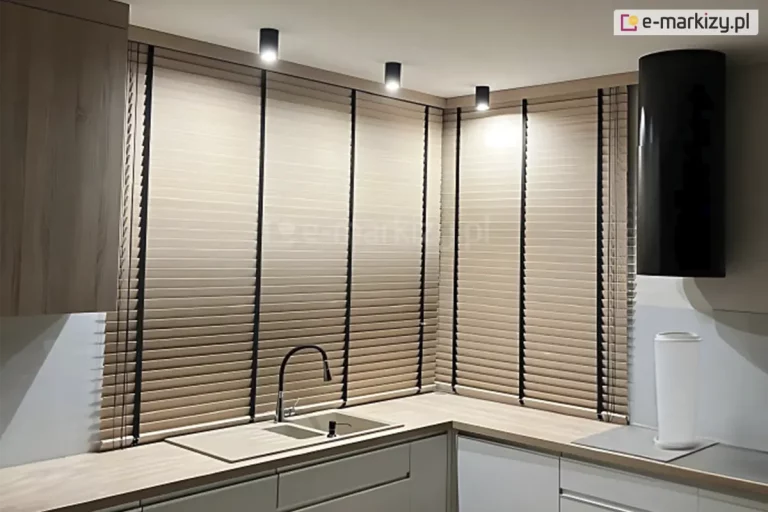 Narożne okno w kuchni osłonięte żaluzjami drewnianymi 50 mm w kolorze tropical beige, z drabinką taśmową w kolorze czarnym, zawieszone pod sufitem z trzema lampami w kształcie tuby, przy zlewie kuchennym wpuszczonym w drewniany blat w kolorze zbliżonym do lamel żaluzji.