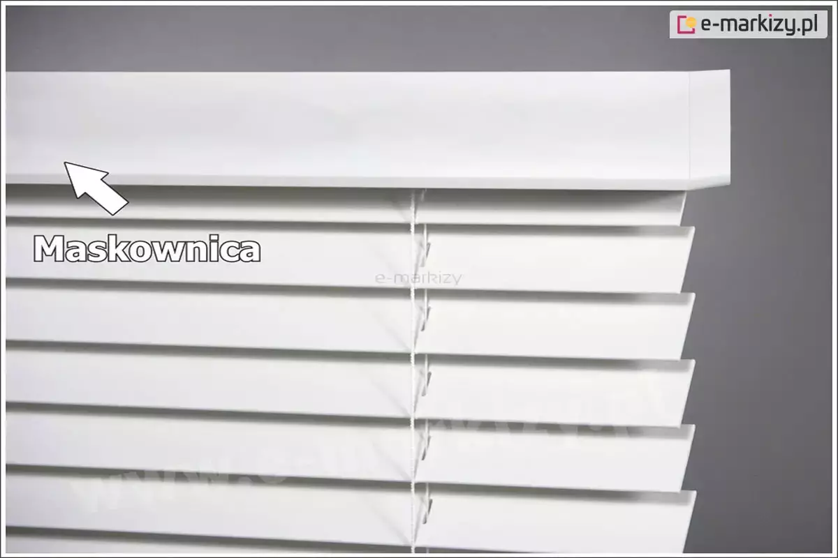 Zbliżenie na górną część białych żaluzji wewnętrznych wykonanych z drewna, skupiające się na estetycznym wykończeniu systemu montażowego. Na pierwszym planie widoczna jest szeroka, biała maskownica o matowej powierzchni, która zakrywa rynnę górną i mechanizm operacyjny. Maskownica ma profil prostokątny, który na prawym końcu został precyzyjnie docięty i zagięty pod kątem, tworząc estetyczne zakończenie (tzw. boczek), idealnie przylegające do bocznej krawędzi żaluzji. Pod maskownicą znajdują się poziome, białe lamele o szerokości 50 mm, ustawione pod lekkim skosem. Przez środek widocznych listewek przebiega cienka, pionowa drabinka sznurkowa w kolorze białym, która utrzymuje lamele w równych odstępach. W lewej części zdjęcia znajduje się biała strzałka skierowana w górę, wskazująca na maskownicę, obok której umieszczono wyraźny, szary napis "Maskownica". Tło za żaluzją jest gładkie i szare, co pozwala na wyraźne wyeksponowanie bieli produktu oraz detali jego konstrukcji. Żaluzje wewnętrzne na zdjęciu prezentują nowoczesny i minimalistyczny design, pasujący do jasnych wnętrz.