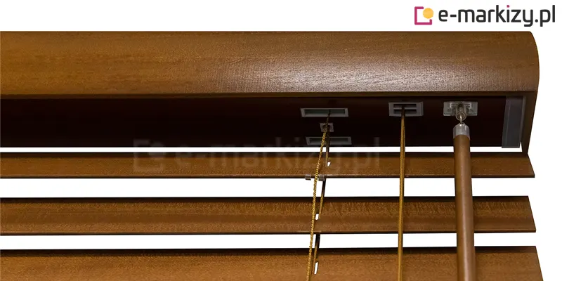 Zdjęcie prezentuje szczegółowe zbliżenie na górną część mechanizmu sterującego, w jaki wyposażone są żaluzje drewniane 25 mm w kolorze średniego, ciepłego brązu o widocznej strukturze słojów. Na pierwszym planie znajduje się górna rynna osłonięta szeroką, drewnianą maskownicą o zaokrąglonym profilu, która w pełni zakrywa elementy montażowe. Bezpośrednio pod rynną widoczne są poziome lamele o szerokości 25 mm, utrzymywane przez system drabinek sznurkowych w kolorze dopasowanym do odcienia drewna. Z mechanizmu wewnątrz rynny wychodzą dwa rodzaje elementów sterujących: po prawej stronie znajduje się sztywny, drewniany drążek (pokrętło) służący do regulacji kąta nachylenia listewek, natomiast obok niego przebiegają cienkie sznurki operacyjne przeznaczone do podnoszenia i opuszczania całej osłony. Sznurki przechodzą przez precyzyjne, prostokątne przelotki w rynnie, co zapewnia płynną pracę systemu. Tło za produktem jest całkowicie białe i neutralne, co pozwala skupić uwagę na detalach wykończenia oraz technicznych aspektach, które wyróżniają te żaluzje drewniane 25 mm.