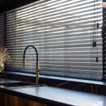 Eleganckie żaluzje bambusowe w kolorze głębokiej, matowej czerni zamontowane na szerokim oknie w luksusowej, nowoczesnej kuchni. Lamele żaluzji są ustawione w pozycji półotwartej, co pozwala na precyzyjną kontrolę wpadającego światła dziennego i tworzy liniowe cienie na blacie roboczym. Konstrukcja opiera się na cienkich drabinkach sznurkowych, które są niemal niewidoczne na tle ciemnych listew. Po prawej stronie zwisają estetyczne sznurki sterujące zakończone minimalistycznymi, cylindrycznymi obciążnikami w kolorze czarnym. Żaluzje bambusowe idealnie komponują się z aranżacją wnętrza, w której dominuje czarny blat z wyraźnym, złotym użyleniem przypominającym marmur oraz stylowa, złoto-czarna bateria kuchenna o wysokiej wylewce. Na blacie pod oknem, po lewej stronie, ustawiono dekoracyjną złotą wazę z suszonymi gałązkami, a po prawej stronie widać fragment ciemnej obudowy okna i nowoczesne akcesoria kuchenne. Całość tworzy prestiżowy i spójny wizualnie klimat, podkreślony przez naturalną teksturę bambusa widoczną pod światło.