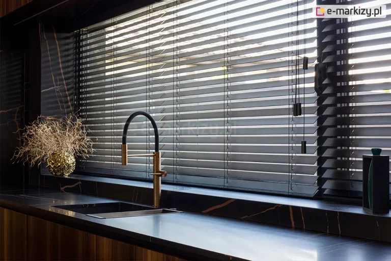 Eleganckie żaluzje bambusowe w kolorze głębokiej, matowej czerni zamontowane na szerokim oknie w luksusowej, nowoczesnej kuchni. Lamele żaluzji są ustawione w pozycji półotwartej, co pozwala na precyzyjną kontrolę wpadającego światła dziennego i tworzy liniowe cienie na blacie roboczym. Konstrukcja opiera się na cienkich drabinkach sznurkowych, które są niemal niewidoczne na tle ciemnych listew. Po prawej stronie zwisają estetyczne sznurki sterujące zakończone minimalistycznymi, cylindrycznymi obciążnikami w kolorze czarnym. Żaluzje bambusowe idealnie komponują się z aranżacją wnętrza, w której dominuje czarny blat z wyraźnym, złotym użyleniem przypominającym marmur oraz stylowa, złoto-czarna bateria kuchenna o wysokiej wylewce. Na blacie pod oknem, po lewej stronie, ustawiono dekoracyjną złotą wazę z suszonymi gałązkami, a po prawej stronie widać fragment ciemnej obudowy okna i nowoczesne akcesoria kuchenne. Całość tworzy prestiżowy i spójny wizualnie klimat, podkreślony przez naturalną teksturę bambusa widoczną pod światło.