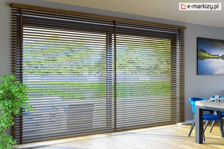 Szerokie żaluzje poziome wykonane z ciemnego drewna, zamontowane na całej powierzchni dużego okna tarasowego w nowoczesnej jadalni. Lamele żaluzji są ustawione w pozycji otwartej, tworząc rytmiczny wzór poziomych linii, przez które prześwituje jasny taras oraz soczysta zielona roślinność w ogrodzie. Całość konstrukcji jest zwieńczona drewnianą maskownicą górną w tym samym ciemnym odcieniu brązu. Pomieszczenie jest jasne, o jasnoszarych ścianach i podłodze z jasnych paneli drewnianych. Po prawej stronie kadru widoczny jest fragment stołu jadalnianego z szarym blatem, przy którym stoją nowoczesne krzesła z intensywnie niebieskim obiciem. Nad stołem, na ścianie, wisi obraz przedstawiający pejzaż z jeziorem, a po lewej stronie znajduje się fragment dużej, zielonej rośliny doniczkowej. Naturalne światło wpadające przez żaluzje tworzy na podłodze pasy światła i cienia, podkreślając przestronność i nowoczesny wystrój wnętrza.