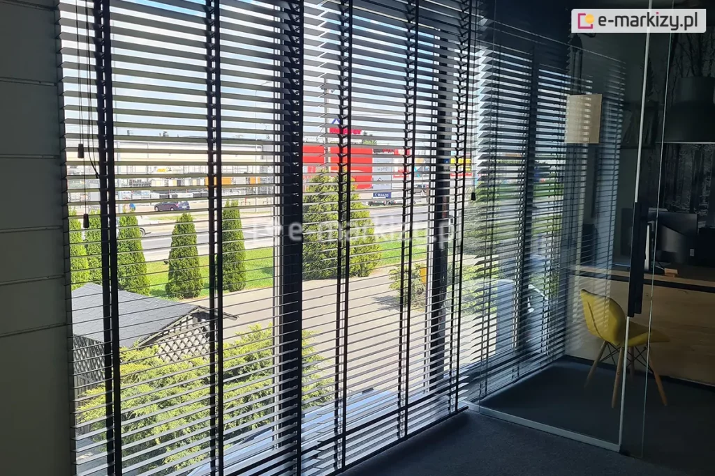 Duże przeszklone okna przesłonięte lamelowymi żaluzjami drewnianymi zaciemniającymi obszerne pomieszczenie