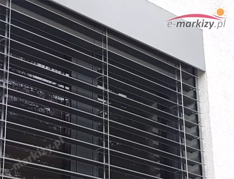 Zewnętrzna żaluzja fasadowa zamontowana na dużym oknie z grafitową ramą aluminiową. Widoczne lamele aluminiowe w układzie poziomym – typ c. Lamele prowadzone są w tekstylnych drabinkach z linkami stabilizującymi oraz dodatkowymi bocznymi prowadnicami wykonanymi z aluminium – te elementy zapewniają większą stabilność całej konstrukcji, szczególnie przy silnym wietrze. Całość osłonięta została estetyczną kasetą górną w kolorze dopasowanym do elewacji – jasnoszarym, o prostym, minimalistycznym kształcie. Kaseta chroni mechanizm podnoszenia żaluzji oraz zwinięte lamele. Okno otoczone jest tynkiem strukturalnym w kolorze białym. Tego typu system osłonowy znajduje zastosowanie zarówno w budownictwie jednorodzinnym, jak i obiektach komercyjnych, oferując zaawansowaną ochronę przeciwsłoneczną, komfort termiczny oraz nowoczesny wygląd elewacji.