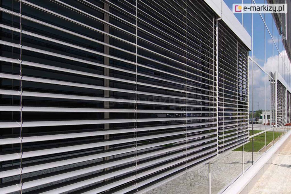 Widok z perspektywy zewnętrznej na nowoczesną elewację budynku biurowego, na której zamontowano system żaluzji fasadowych. Szerokie, poziome lamele w kolorze srebrnym lub jasnoszarym są ustawione pod kątem, co pozwala na regulację wpadającego światła przy jednoczesnym ograniczeniu nagrzewania się wnętrza. Konstrukcja żaluzji opiera się na pionowych prowadnicach listwowych, które stabilizują poszczególne pióra systemu. Żaluzje zamocowane są wzdłuż dużych przeszkleń odbijających błękitne niebo oraz białe chmury. Na pierwszym planie widoczny jest detal ułożenia lameli oraz system ich zawieszenia, natomiast w tle widać dalszą część fasady z kolejnymi modułami osłon, fragment zielonego trawnika oraz brukowany chodnik przy podstawie obiektu.