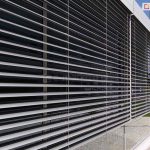 Widok z perspektywy zewnętrznej na nowoczesną elewację budynku biurowego, na której zamontowano system żaluzji fasadowych. Szerokie, poziome lamele w kolorze srebrnym lub jasnoszarym są ustawione pod kątem, co pozwala na regulację wpadającego światła przy jednoczesnym ograniczeniu nagrzewania się wnętrza. Konstrukcja żaluzji opiera się na pionowych prowadnicach listwowych, które stabilizują poszczególne pióra systemu. Żaluzje zamocowane są wzdłuż dużych przeszkleń odbijających błękitne niebo oraz białe chmury. Na pierwszym planie widoczny jest detal ułożenia lameli oraz system ich zawieszenia, natomiast w tle widać dalszą część fasady z kolejnymi modułami osłon, fragment zielonego trawnika oraz brukowany chodnik przy podstawie obiektu.