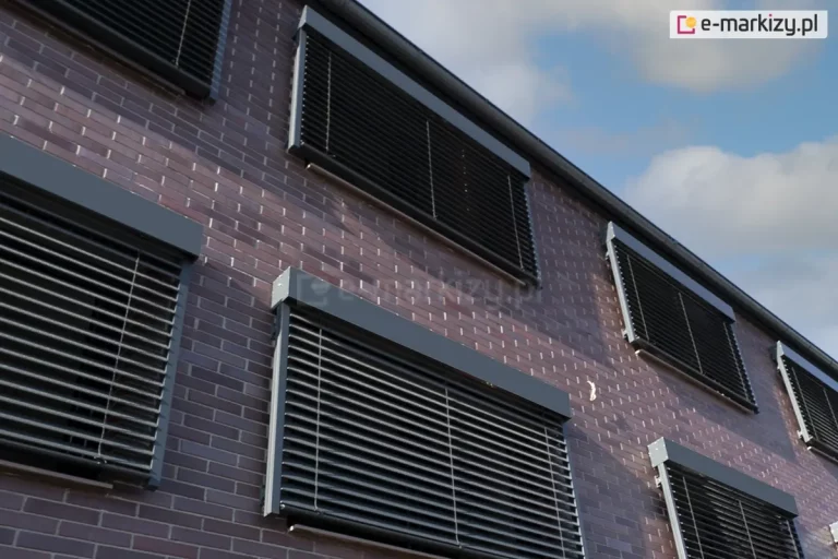 Ceglany budynek z oknami osłoniętymi żaluzjami fasadowymi w kasecie prostokątnej model Cube Selt w kolorze czarnym. Nad budynkiem widoczne niebieskie niebo przesłonięte częściowo chmurami.