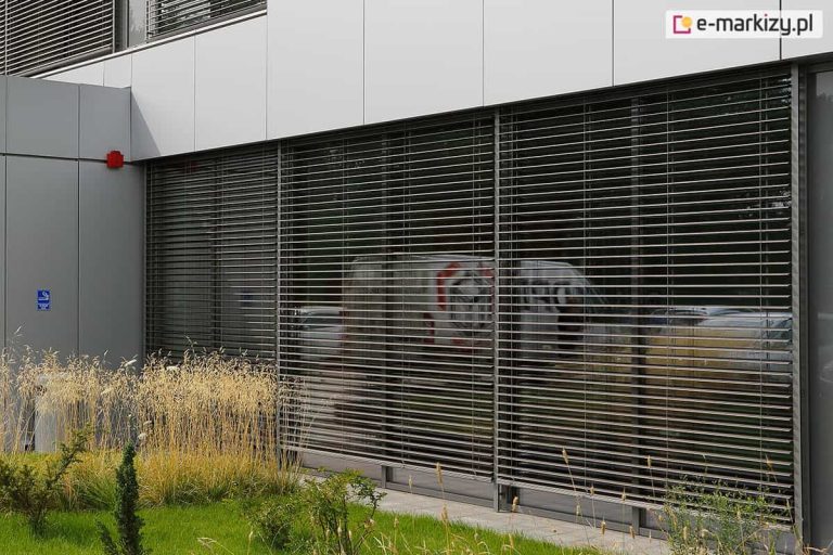 Duża przeszklona ściana na parterze budynku produkcyjnego lub biurowego, przesłonięta połączonymi modułowo żaluzjami fasadowymi BOX z ukrytymi pod elewacją skrzynkami. Częściowo uchylone lamele pozwalają dostrzec w odbiciu szyby zaparkowane w pobliżu auta. Przed fasadą zielony trawnik z ozdobnymi roślinami.