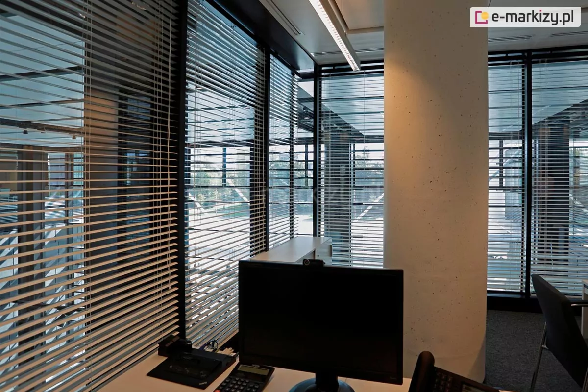 Widok z perspektywy stanowiska pracy na narożne przeszklenie biurowe wyposażone w system żaluzji fasadowych C50 Slim Selt. Aluminiowe lamele w kolorze srebrnym są ustawione w pozycji otwartej, co pozwala na precyzyjne rozproszenie ostrego światła słonecznego przy jednoczesnym zachowaniu wglądu na architekturę sąsiednich budynków i infrastrukturę miejską. Wnętrze charakteryzuje się nowoczesnym, surowym stylem z widocznym betonowym słupem konstrukcyjnym oraz liniowym oświetleniem sufitowym. Na pierwszym planie znajduje się monitor komputerowy oraz telefon stacjonarny, co obrazuje skuteczność żaluzji w eliminowaniu uciążliwych odblasków na ekranach w mocno przeszklonych pomieszczeniach typu open space. System żaluzji montowany jest do ciemnych profili okiennych, co podkreśla techniczny i profesjonalny charakter aranżacji.