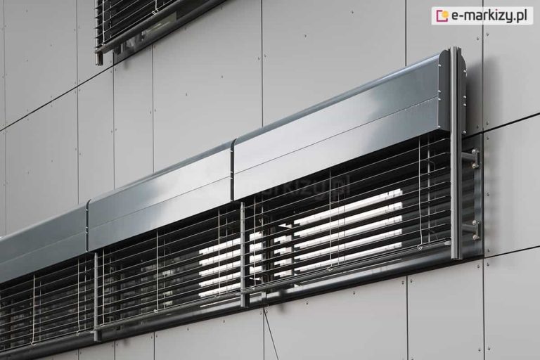 Zbliżenie na detale techniczne zewnętrznych żaluzji fasadowych C80 Oval Selt zamontowanych na nowoczesnym budynku biurowym. Zdjęcie przedstawia system osłon okiennych w kolorze antracytowym, zainstalowany na elewacji wykonanej z jasnoszarych paneli kompozytowych. Widoczna jest solidna kaseta osłonowa o półokrągłym profilu, chroniąca mechanizm zwijający. Poziome lamele żaluzji są częściowo otwarte, co pozwala na regulację dopływu światła do wnętrza. Konstrukcja zawiera pionowe prowadnice listwowe oraz stalowe uchwyty montażowe przytwierdzone bezpośrednio do elewacji za pomocą widocznych śrub. Precyzyjne spasowanie modułów żaluzji wskazuje na systemowy montaż na szerokim przeszkleniu. Powierzchnia paneli elewacyjnych jest matowa, kontrastując z lekko połyskującym wykończeniem aluminiowych elementów żaluzji. Całość prezentuje estetyczne i funkcjonalne rozwiązanie z zakresu nowoczesnej architektury, mające na celu poprawę efektywności energetycznej budynku.