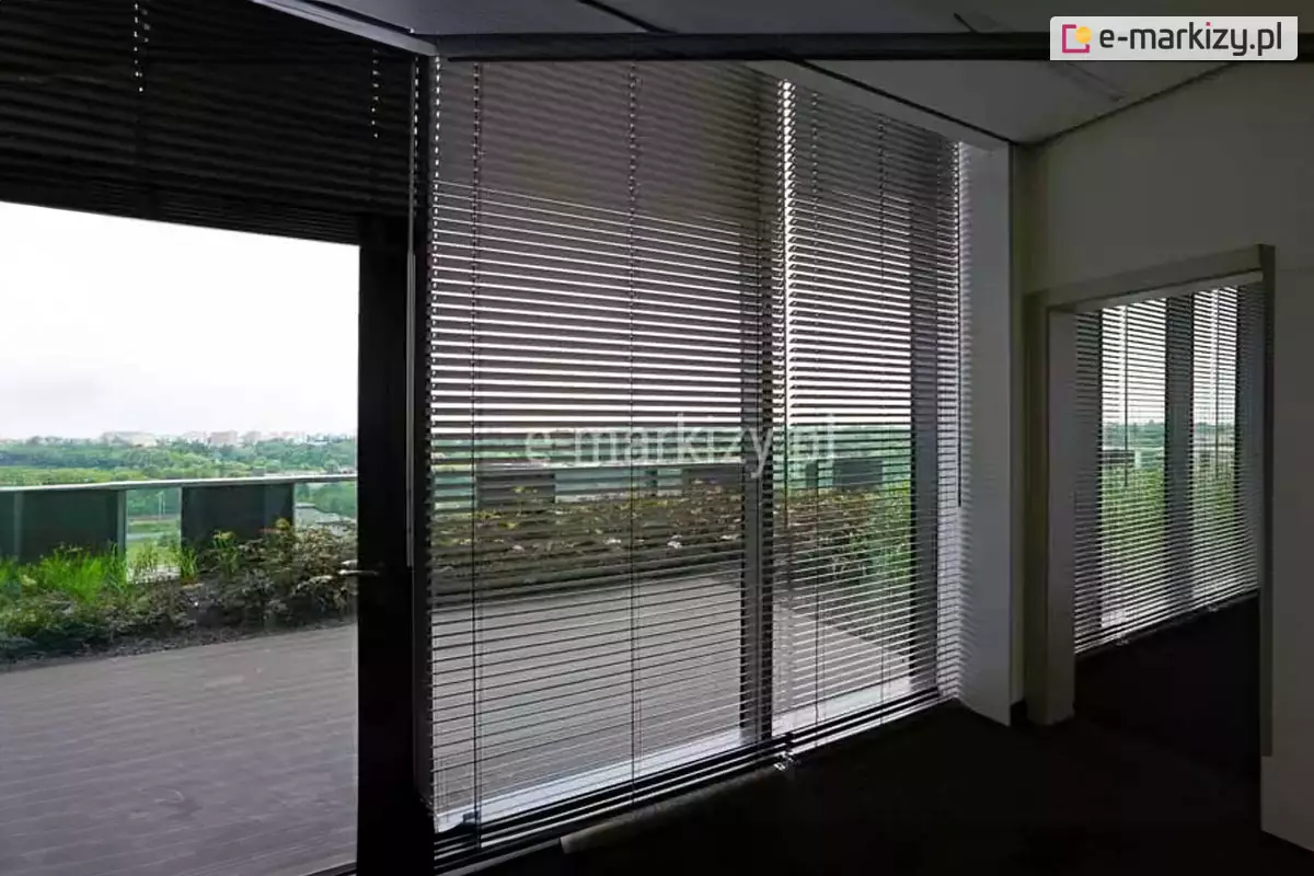 Widok z wnętrza nowoczesnego biura na system wewnętrznych żaluzji fasadowych C50 zamontowanych na wielkoformatowych przeszkleniach tarasowych. Srebrne, aluminiowe lamele o wysokim połysku są ustawione w pozycji otwartej, co pozwala na precyzyjne filtrowanie światła dziennego i redukcję nagrzewania pomieszczenia przy zachowaniu widoczności na zewnętrzny taras widokowy. Przez żaluzje widoczna jest balustrada tarasu, niska zieleń ozdobna oraz rozległa panorama miasta w tle. Konstrukcja osłon obejmuje system prowadzenia bocznego, który stabilizuje żaluzje przy otwieraniu drzwi balkonowych. Ciemna podłoga biura kontrastuje z jasnym wykończeniem sufitu i ścian, podkreślając techniczny, a zarazem elegancki charakter zastosowanego systemu zaciemnienia fasadowego.