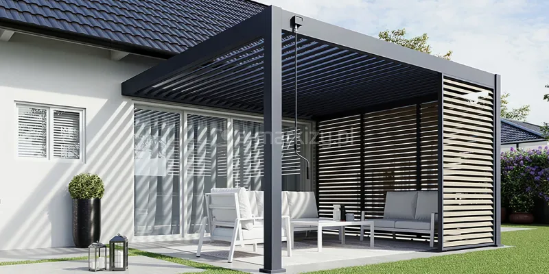Parterowy dom z antracytowym dwuspadowym dachem oraz białą elewacją, do której przymocowano pergolę lamelową Rubio Soft w kolorze zbliżonym do pokrycia dachu. Zadaszenie pergoli wyposażone jest w ruchome lamele dachowe, które na zdjęciu są częściowo otwarte, rzucając geometryczne cienie na jasną ścianę budynku oraz przeszklone drzwi tarasowe. Konstrukcja posiada zintegrowane przesłony boczne w formie paneli żaluzjowych, wykonanych z jasnego drewna, które osłaniają prawą stronę tarasu, zapewniając prywatność i ochronę przed słońcem. Pod pergolą znajduje się komfortowa strefa wypoczynkowa wyposażona w białe meble ogrodowe z grubymi, jasnymi poduchami, w tym kanapy oraz fotel ustawione wokół niskiego stolika kawowego. Taras jest wyłożony jasnymi płytami, a obok mebli widoczne są dekoracyjne latarnie ogrodowe oraz wysoka, czarna donica z przystrzyżonym, zielonym krzewem o kulistym kształcie. W tle po prawej stronie widać fragment zadbanego ogrodu z zielonym trawnikiem oraz fioletowymi kwiatami. Całość prezentuje minimalistyczny i funkcjonalny design zewnętrznej przestrzeni relaksu w pełnym świetle dziennym.
