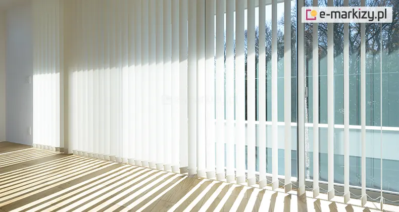 Zdjęcie przedstawia nowoczesne wnętrze jasnego pomieszczenia z dużymi przeszkleniami, na których zamontowano białe żaluzje pionowe zwane verticalami. Lamele żaluzji są ustawione pod kątem, co pozwala na częściowe przenikanie naturalnego światła słonecznego do środka. Promienie słońca wpadające przez szczeliny między pasami materiału tworzą na jasnej, drewnianej podłodze regularny wzór równoległych, świetlistych pasów naprzemiennie z cieniami. Okno zajmuje niemal całą wysokość ściany, a za szybami widoczne są korony drzew pozbawione liści, co sugeruje okres jesienno-zimowy, oraz fragmenty błękitnego nieba. System osłon okiennych składa się z wielu pionowych pasów tkaniny zawieszonych na górnej szynie, które u dołu są połączone białym łańcuszkiem stabilizującym. Wnętrze jest minimalistyczne, z białymi ścianami, co potęguje wrażenie przestronności i czystości. Światło jest bardzo intensywne, powodując lekkie prześwietlenie w centralnej części kadru, gdzie promienie bezpośrednio uderzają w krawędzie lameli.