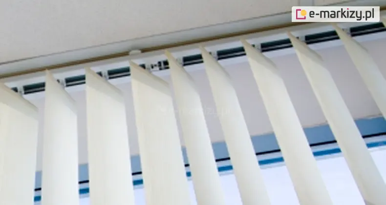 Zdjęcie prezentuje zbliżenie na górny mechanizm montażowy żaluzji pionowych, znanych jako verticale, zamontowanych bezpośrednio pod sufitem. Widoczna jest biała, aluminiowa szyna prowadząca, w której osadzone są ruchome wózki z haczykami trzymającymi poszczególne lamele. Pasy tkaniny w kolorze kremowym lub bardzo jasnym beżu są zawieszone pionowo i ustawione pod skosem względem płaszczyzny okna, co ukazuje technikę regulacji kąta nachylenia materiału. Każda lamelka ma precyzyjnie wykończoną górną krawędź wsuniętą w uchwyt mocujący. W tle, za rozsuniętymi pasami materiału, widoczny jest fragment ramy okiennej oraz jasne, rozproszone światło dzienne wpadające z zewnątrz. Górna część kadru obejmuje fragment sufitu o delikatnej strukturze, do którego przylega szyna verticali. Perspektywa zdjęcia jest skierowana od dołu ku górze, co pozwala dokładnie przyjrzeć się elementom konstrukcyjnym systemu osłonowego oraz sposobowi, w jaki poszczególne pasy tkaniny wychodzą z mechanizmu jezdnego.