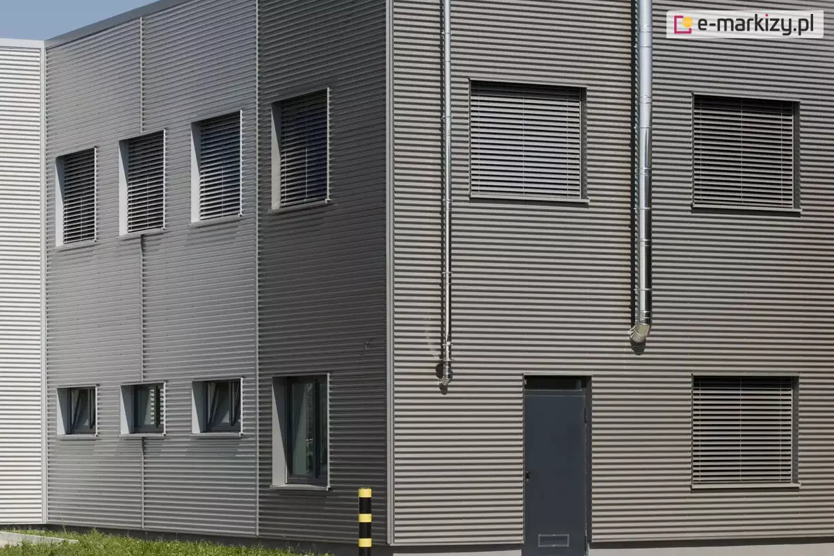 Narożnik nowoczesnego, dwukondygnacyjnego budynku o elewacji z szarej, poziomej blachy trapezowej. Na obu widocznych ścianach zamontowane są zewnętrzne żaluzje fasadowe z poziomymi aluminiowymi lamelami w kolorze srebrnym. Żaluzje osłaniają wszystkie okna na piętrze – cztery od strony lewej oraz trzy od frontu. Dodatkowo, na parterze widoczne są trzy okna bez żaluzji po lewej stronie oraz jedno większe po prawej stronie, także bez osłony. Po lewej stronie narożnika znajduje się słupek ochronny w kolorze żółto-czarnym, ustawiony przed ścianą budynku. Drzwi techniczne w kolorze grafitowym umieszczone są pośrodku prawej ściany. Całość sugeruje obiekt o przeznaczeniu przemysłowym lub biurowym. Podtynkowe żaluzje i ich lamele współgrają z poziomymi pasami elewacji tworząc interesujący efekt.