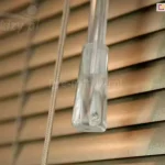 Zbliżenie na detale techniczne sterowania, jakie posiadają żaluzje aluminiowe 16 mm z interesującą teksturą w ciepłym odcieniu brązu. Lamele mają subtelny rysunek wzorów, a ich krawędzie są gładko wykończone i lekko zaokrąglone. Na pierwszym planie widoczny jest pionowy, akrylowy pręt o okrągłym przekroju, służący do precyzyjnego obracania listew i regulacji kąta wpadającego światła. Obok pręta znajdują się białe sznurki operacyjne, które są spięte przezroczystym, plastikowym dzwonkiem o stożkowym kształcie, ułatwiającym chwytanie i podnoszenie całej osłony. W tle widać kolejne poziome segmenty żaluzji ułożone kaskadowo, co tworzy efekt głębi. Oświetlenie pada z lewej strony, akcentując matową powierzchnię aluminium oraz detale mechanizmu sznurkowego. Całość kompozycji skupia się na funkcjonalności i estetyce wykończenia, podkreślając wysoką jakość elementów, z których składa się żaluzja aluminiowa 16 mm.