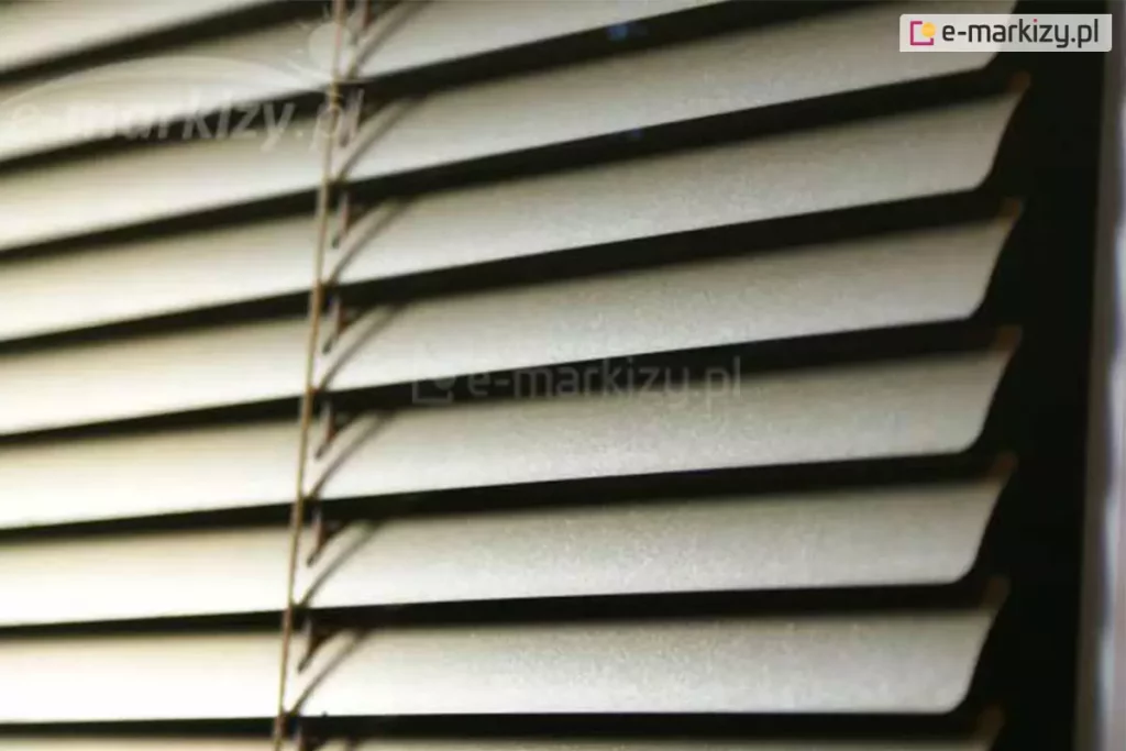 Żaluzja pozioma aluminiowa z wąskimi lamelami w kolorze srebrnym oraz drabinką sznurkową w kolorze zbliżonym do brązu zamocowana na oknie, za którym panuje ciemność.