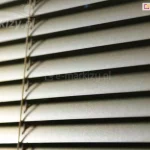 Żaluzja pozioma aluminiowa z wąskimi lamelami w kolorze srebrnym oraz drabinką sznurkową w kolorze zbliżonym do brązu zamocowana na oknie, za którym panuje ciemność.