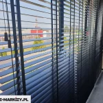Zdjęcie przedstawia nowoczesne wnętrze biurowe lub mieszkalne z szerokim przeszkleniem, na którym zamontowano żaluzje drewniane 25 mm w kolorze antracytowym lub głębokiej czerni. Żaluzje są ustawione w pozycji poziomej, z lamelami lekko nachylonymi, co pozwala na kontrolowany dopływ światła dziennego do pomieszczenia. Konstrukcja opiera się na cienkich lamelach o szerokości 25 mm, które są połączone systemem drabinek sznurkowych w tym samym kolorze co drewno. Po lewej stronie widoczne są zwisające sznurki służące do regulacji wysokości oraz kąta nachylenia listewek, zakończone estetycznymi, podłużnymi obciążnikami. Przez szczeliny między lamelami widać panoramę zewnętrzną, obejmującą parking, drogę, niską zabudowę komercyjną oraz zielone drzewa iglaste i liściaste pod jasnym, bezchmurnym niebem. Wewnątrz pomieszczenia, po prawej stronie kadru, znajduje się fragment biurka o jasnym, drewnianym blacie oraz żółte, nowoczesne krzesło na drewnianych nogach z metalowymi wzmocnieniami. Podłoga wyłożona jest szarą wykładziną o wyraźnej strukturze. Światło padające z okna tworzy na powierzchni lameli subtelne przejścia tonalne, podkreślając matowe wykończenie materiału, z którego wykonano żaluzje drewniane 25 mm.