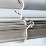Zdjęcie prezentuje skrajne zbliżenie na dolną sekcję żaluzji drewnianych w kolorze białym lub bardzo jasnym popielu, koncentrując się na detalach technicznych wykończenia. Na pierwszym planie widoczny jest dolny profil dociążający, który jest grubszy od pozostałych lameli i posiada zaokrąglone krawędzie. Przez profil ten przechodzi pionowy, biały sznurek drabinkowy, który jest precyzyjnie zawiązany i zabezpieczony od spodu półkolistą, białą zatyczką maskującą węzeł. Powyżej profilu dociążającego widoczne są pozostałe poziome lamele, ułożone w stosie, co sugeruje, że żaluzje drewniane są w trakcie podnoszenia. Przez podłużne otwory w górnych lamelach przechodzą fragmenty sznurka operacyjnego w kolorze brązowym, co tworzy techniczny detal konstrukcyjny. Ostrość zdjęcia jest ustawiona na węzeł sznurka przy dolnej belce, podczas gdy tło pozostaje jasne i rozmyte, co podkreśla czystość formy i precyzję montażu elementów sterujących.