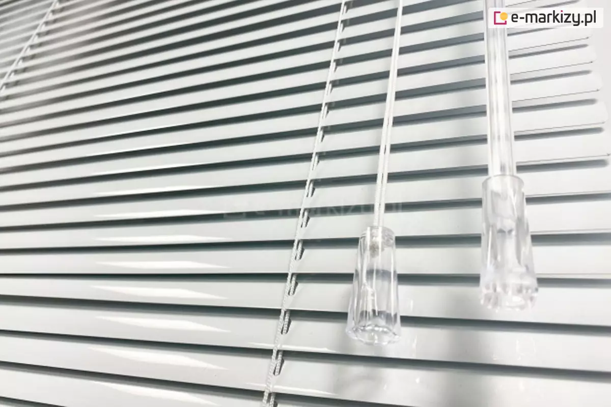 Białe żaluzje aluminiowe z lamelami 25mm o dużym połysku z białą drabinką sznurkową. Po prawej stronie widoczny sznurek do sterowania zakończony lexanowym dzwonkiem oraz pokrętło wykonana z lexanu do zmiany kąta nachylenia lameli.