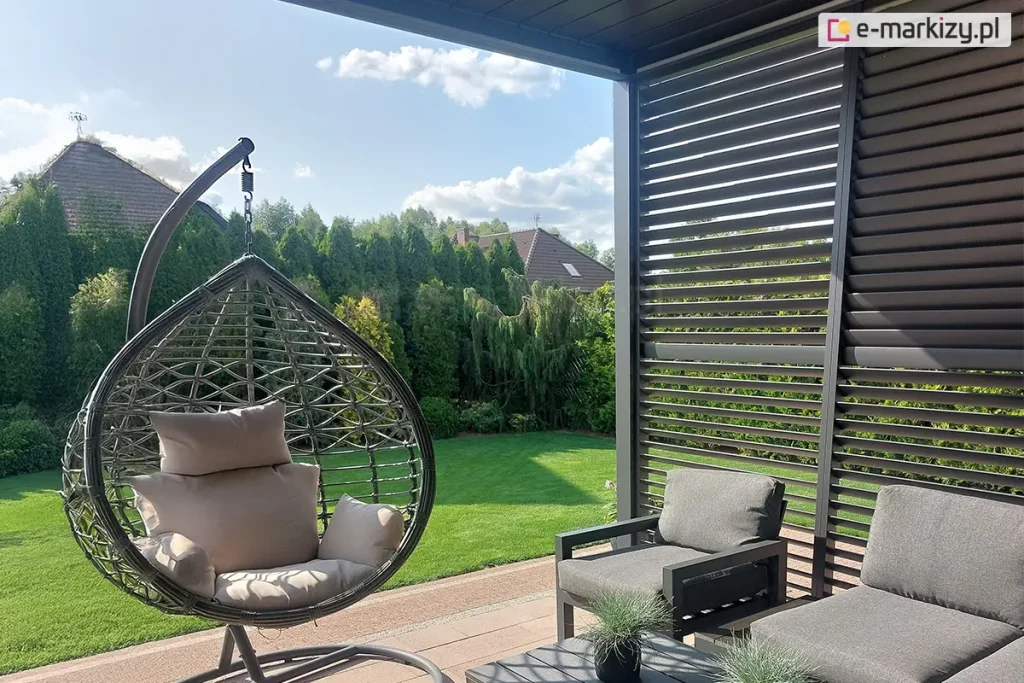 Zdjęcie przedstawia aranżację nowoczesnego tarasu wyposażonego w pergolę lamelową oraz meble ogrodowe, osadzoną w otoczeniu zadbanej zieleni. Po prawej stronie kadru widoczna jest boczna ściana pergoli w kolorze antracytowym, którą tworzą poziome, ruchome lamele ustawione pod kątem, co pozwala na prześwitywanie naturalnego światła dziennego. Konstrukcja pergoli graniczy z krawędzią dachu widoczną u góry zdjęcia. Na pierwszym planie po lewej stronie znajduje się wiszący fotel ogrodowy typu kokon w kolorze ciemnoszarym, zawieszony na solidnym metalowym stelażu, wyposażony w jasnoszare, miękkie poduszki siedziskowe i oparciowe. W centralnej części i po prawej stronie umieszczono zestaw wypoczynkowy składający się z dwóch foteli o prostej, geometrycznej ramie w kolorze ciemnym, z grubymi grafitowymi poduchami. Przed meblami stoi niski stolik kawowy, na którym ustawiono dwie małe doniczki z ozdobnymi trawami. Podłoże tarasu wyłożone jest jasnobrązową kostką brukową lub płytami tarasowymi. Tło kompozycji stanowi intensywnie zielony, równo przystrzyżony trawnik, za którym rozciąga się gęsty żywopłot z wysokich tuj oraz inne ozdobne krzewy i drzewa iglaste. W oddali, ponad roślinnością, widoczne są dachy domów jednorodzinnych kryte brązową dachówką, a nad nimi błękitne niebo z lekkimi, rozproszonymi chmurami. Światło słoneczne pada z lewej strony, tworząc wyraźne cienie na meblach i nawierzchni tarasu.