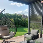 Zdjęcie przedstawia aranżację nowoczesnego tarasu wyposażonego w pergolę lamelową oraz meble ogrodowe, osadzoną w otoczeniu zadbanej zieleni. Po prawej stronie kadru widoczna jest boczna ściana pergoli w kolorze antracytowym, którą tworzą poziome, ruchome lamele ustawione pod kątem, co pozwala na prześwitywanie naturalnego światła dziennego. Konstrukcja pergoli graniczy z krawędzią dachu widoczną u góry zdjęcia. Na pierwszym planie po lewej stronie znajduje się wiszący fotel ogrodowy typu kokon w kolorze ciemnoszarym, zawieszony na solidnym metalowym stelażu, wyposażony w jasnoszare, miękkie poduszki siedziskowe i oparciowe. W centralnej części i po prawej stronie umieszczono zestaw wypoczynkowy składający się z dwóch foteli o prostej, geometrycznej ramie w kolorze ciemnym, z grubymi grafitowymi poduchami. Przed meblami stoi niski stolik kawowy, na którym ustawiono dwie małe doniczki z ozdobnymi trawami. Podłoże tarasu wyłożone jest jasnobrązową kostką brukową lub płytami tarasowymi. Tło kompozycji stanowi intensywnie zielony, równo przystrzyżony trawnik, za którym rozciąga się gęsty żywopłot z wysokich tuj oraz inne ozdobne krzewy i drzewa iglaste. W oddali, ponad roślinnością, widoczne są dachy domów jednorodzinnych kryte brązową dachówką, a nad nimi błękitne niebo z lekkimi, rozproszonymi chmurami. Światło słoneczne pada z lewej strony, tworząc wyraźne cienie na meblach i nawierzchni tarasu.