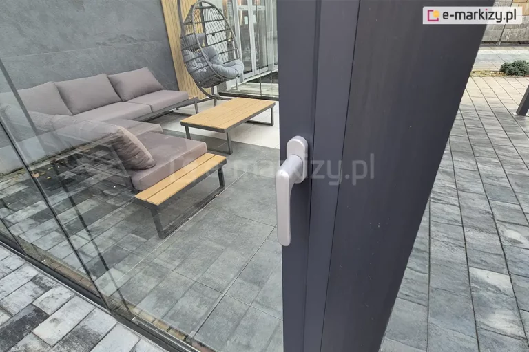 Zbliżenie na system zamknięcia drzwi przesuwnych Auris z widoczną klamką. Na zdjęciu widać profil skrzydła drzwiowego, mechanizm ryglowania oraz aluminiową klamkę zamontowaną po zewnętrznej stronie prowadnicy. Za szklanymi drzwiami, na wyłożonym płytkami betonowymi tarasie ustawiono fotel typu kokon oraz zestaw mebli tarasowych z brązowymi poduszkami i niskim stolikiem z drewnianym blatem.