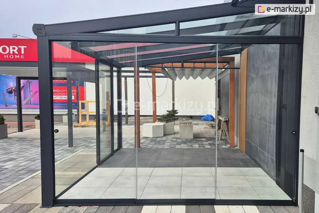 Zdjęcie przedstawia nowoczesną konstrukcję aluminiową z rozsuwanym przeszkleniem – zabudowę tarasową Terano, zlokalizowaną na ekspozycji zewnętrznej. Front i boczna ściana wykonane są z przezroczystych paneli szklanych bez ramek Miros, osadzonych w czarnej aluminiowej ramie. Dach zadaszenia wykonany ze szkła bezpiecznego. W tle widoczna pergola drewniana z dachem rzymskim tkaninowym Supro. W tle ekspozycja sklepu z billboardem reklamowym oraz elementami aranżacyjnymi, takimi jak donice, ławki, drabina i płyty tarasowe.