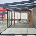 Zdjęcie przedstawia nowoczesną konstrukcję aluminiową z rozsuwanym przeszkleniem – zabudowę tarasową Terano, zlokalizowaną na ekspozycji zewnętrznej. Front i boczna ściana wykonane są z przezroczystych paneli szklanych bez ramek Miros, osadzonych w czarnej aluminiowej ramie. Dach zadaszenia wykonany ze szkła bezpiecznego. W tle widoczna pergola drewniana z dachem rzymskim tkaninowym Supro. W tle ekspozycja sklepu z billboardem reklamowym oraz elementami aranżacyjnymi, takimi jak donice, ławki, drabina i płyty tarasowe.