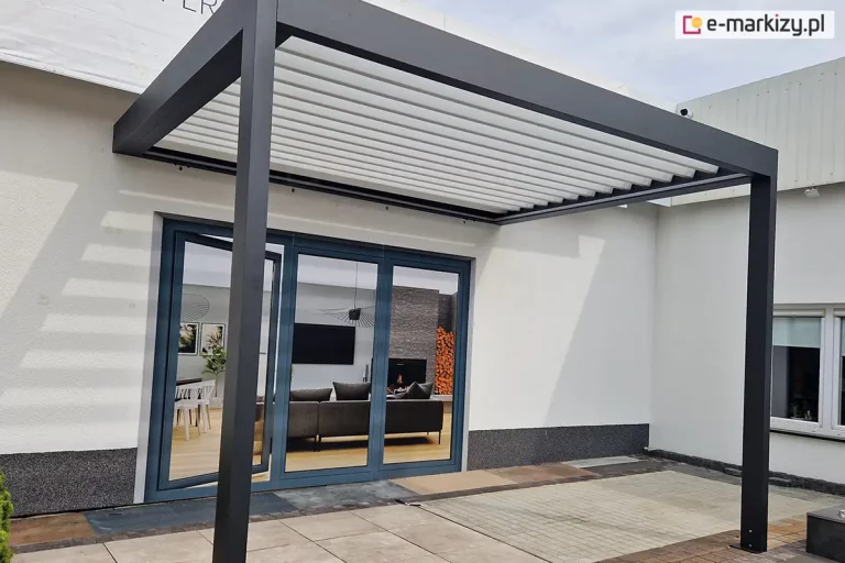 Aluminiowa pergola tarasowa SB400 w kolorze ciemnego grafitu, przymocowana do białej ściany budynku z ciemnoszarym cokołem. Zadaszenie bioklimatyczne z uchylonymi białymi lamelami wspiera się na dwóch nogach o kwadratowym przekroju, zakotwiczonych w podłożu wykonanym z jasnych płyt tarasowych i kostki brukowej. Przeszklone drzwi przesuwne pod pergolą ukazują odbicie nowocześnie urządzonego salonu z szarymi meblami, stołem z krzesłami oraz dekoracyjnym stosem drewna przy kominku. Na elewacji po lewej stronie rzucany jest charakterystyczny liniowy cień ramy pergoli. Po prawej stronie widoczny jest fragment białej ściany z małym oknem i wystającą kamerą monitoringu przy krawędzi dachu. Całość prezentowana jest na tle jasnego, jesiennego nieba.